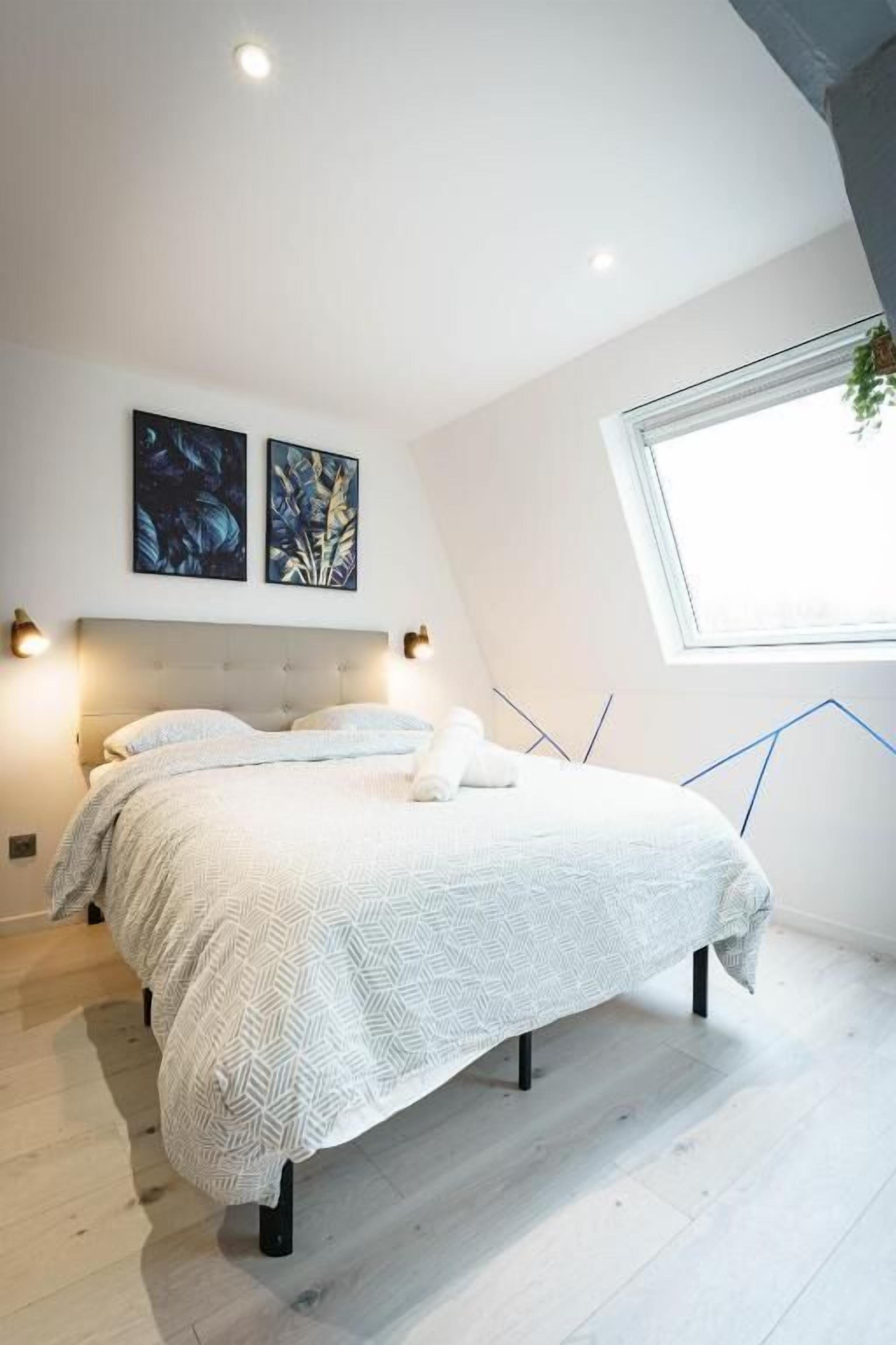 A bedroom with a bed, two wall-mounted light fixtures, a window, and two framed artworks.