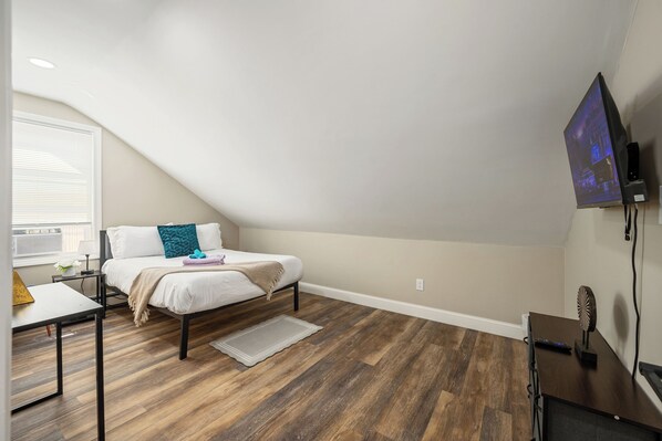 A bedroom with a sloped ceiling, a bed, a desk, and a TV mounted on the wall.