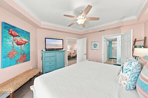 Ein Schlafzimmer mit einem Bett, einem Deckenventilator, einem Flamingo-Gemälde, einem Fernseher und einem Schminktisch.