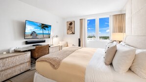A hotel room with a bed, a TV, a desk, a chair, a window, and a painting. - 1-bedroom hotel suites in amazing Miami Beach with fitness room (Miami Beach)