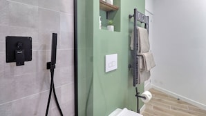 Casa de banho com cabine de chuveiro em vidro, porta-toalhas com toalhas, rolo de papel higiénico e uma pequena prateleira com uma planta.