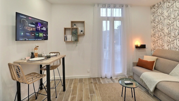 Uma sala de estar com sofá, mesa de jantar com cadeiras, televisão na parede e janela com cortinas.