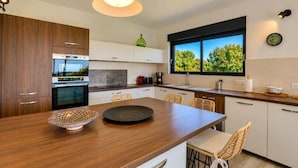 A modern kitchen with wooden cabinets, a large island, and a window with a view of trees. - Villa des Pins (Ventabren)