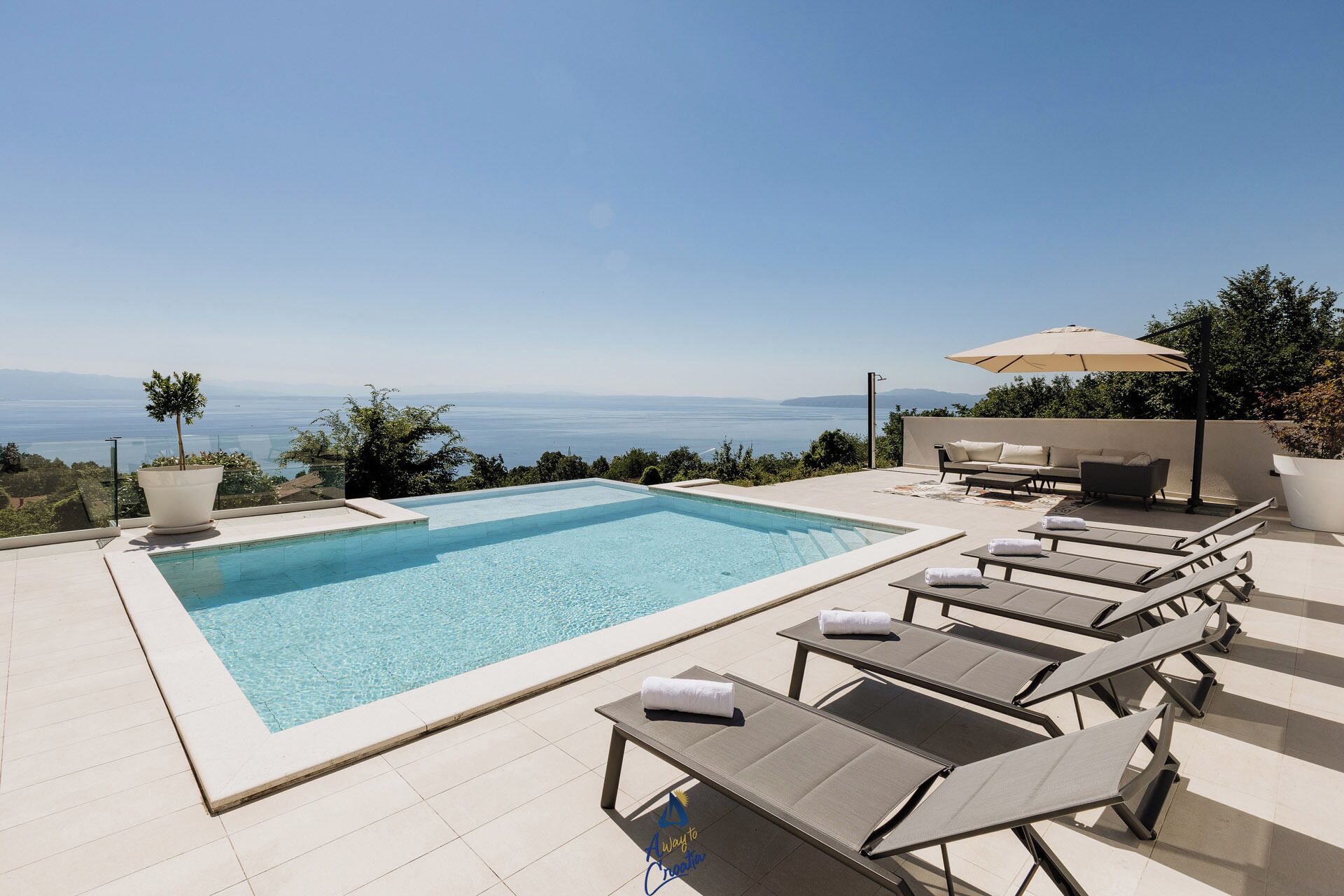 A pool with lounge chairs and a sofa, a potted plant, and a view of the sea.
