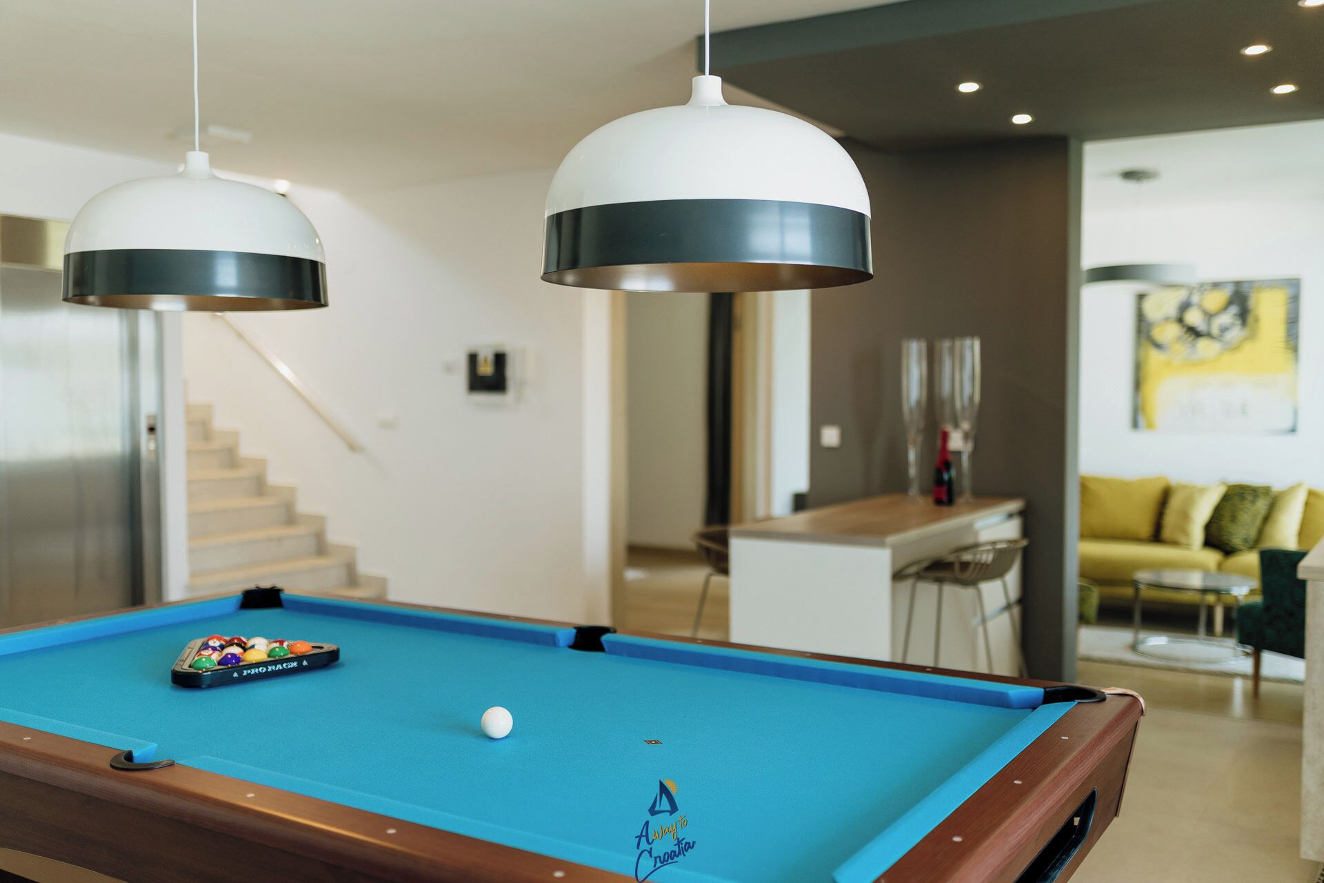 Una mesa de billar con bola blanca y un juego de bolas de pool, dos lámparas colgantes, una escalera y un sofá amarillo al fondo.