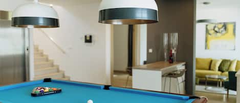 Una mesa de billar con bola blanca y un juego de bolas de pool, dos lámparas colgantes, una escalera y un sofá amarillo al fondo.