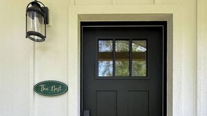 Une porte d’entrée sombre avec une petite fenêtre, une enseigne verte portant l’inscription « The Nest », et une lanterne fixée au mur.