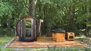Une structure en bois comprenant une grande pièce en forme de tonneau, une plus petite pièce également en forme de tonneau et une pièce carrée dotée d’une fenêtre, le tout sur une terrasse en bois entourée d’arbres.
