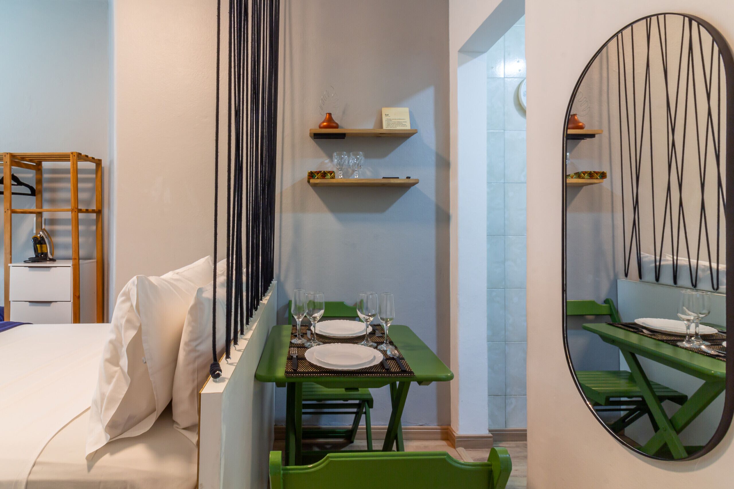 A hotel room with a bed, a small table with a green chair, a mirror, and shelves with items.