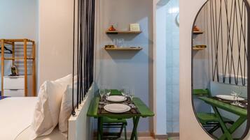 A hotel room with a bed, a small table with a green chair, a mirror, and shelves with items.