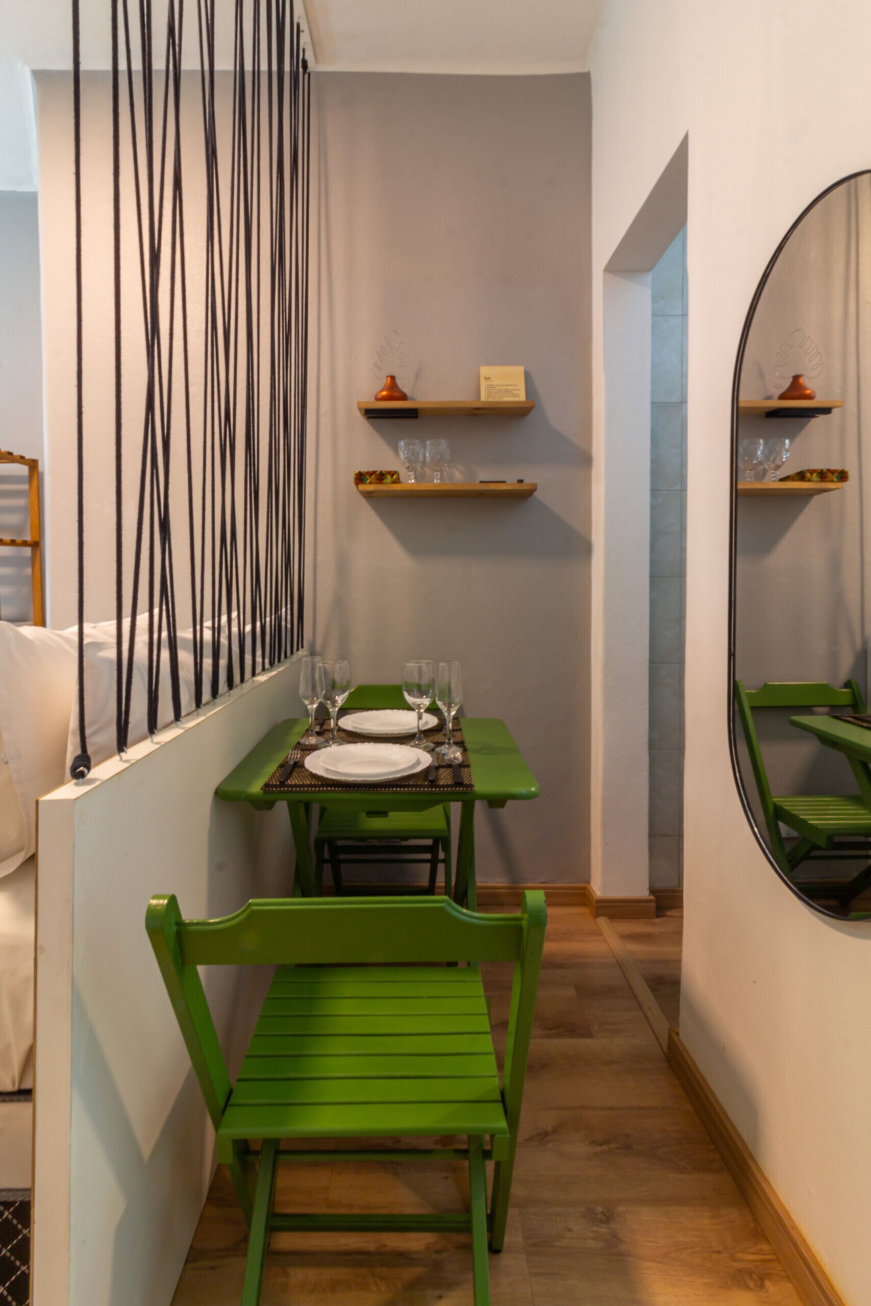 Un couloir étroit mène à un coin repas où se trouvent une table et des chaises vertes, un miroir et des étagères murales ornées d’objets décoratifs.