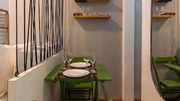 A narrow hallway with a dining area featuring a green table and chairs, a mirror, and wall-mounted shelves with decorative items.