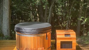 Eine Holzhütte als Sauna mit einer Bank und einer Bedieneinheit, auf einer Holzterrasse in einem Wald gelegen.