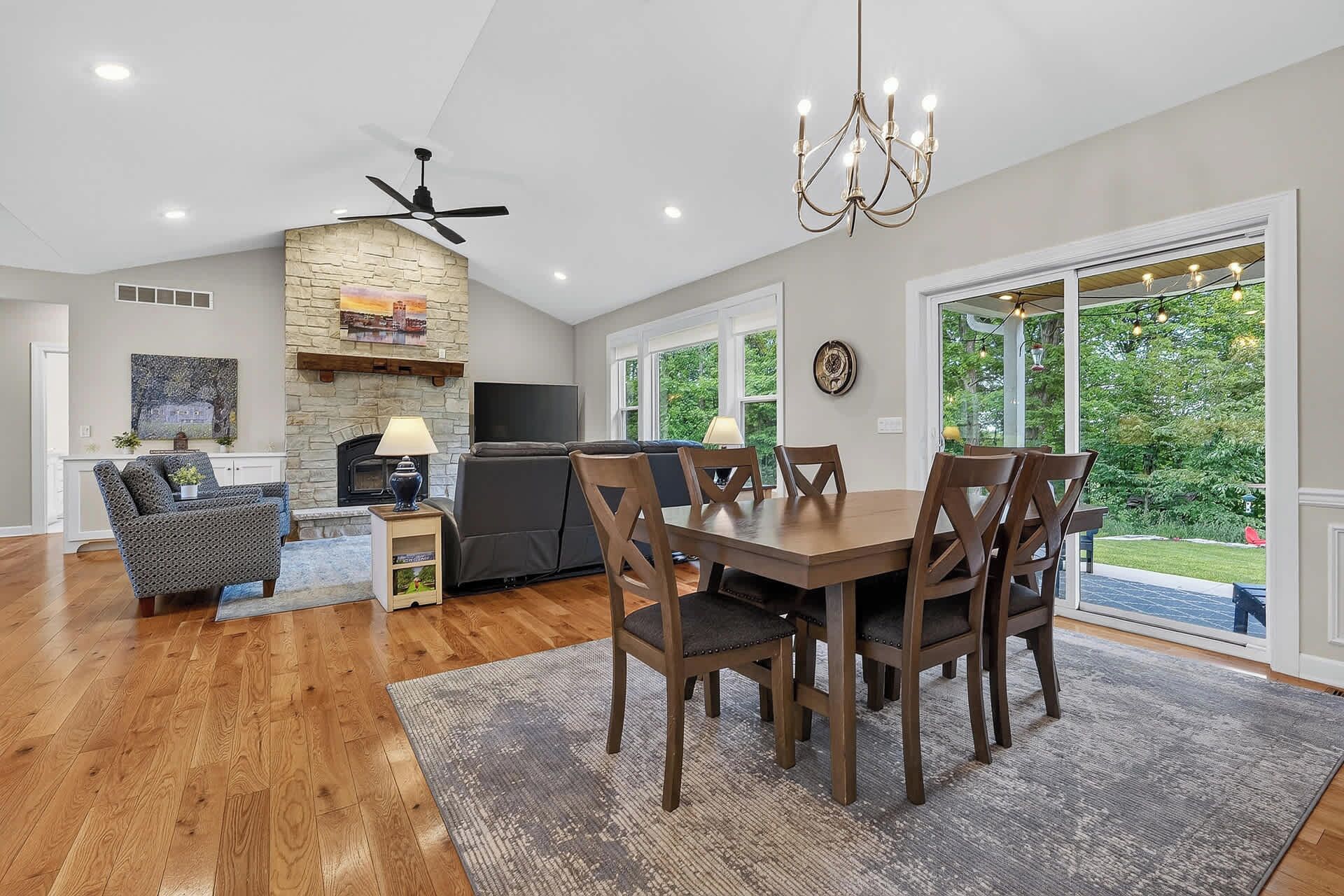 A spacious living room with a fireplace, a dining area with wooden chairs, a ceiling fan, and large windows offering a view of the outdoors.
