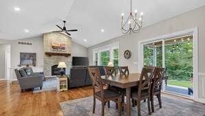 Una amplia sala de estar con chimenea, un comedor con sillas de madera, un ventilador de techo y grandes ventanales con vistas al exterior.