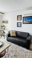 Uma sala de estar moderna com um sofá de couro preto, uma escrivaninha com um computador, uma cadeira e uma pequena planta. Há quadros nas paredes e uma janela grande.