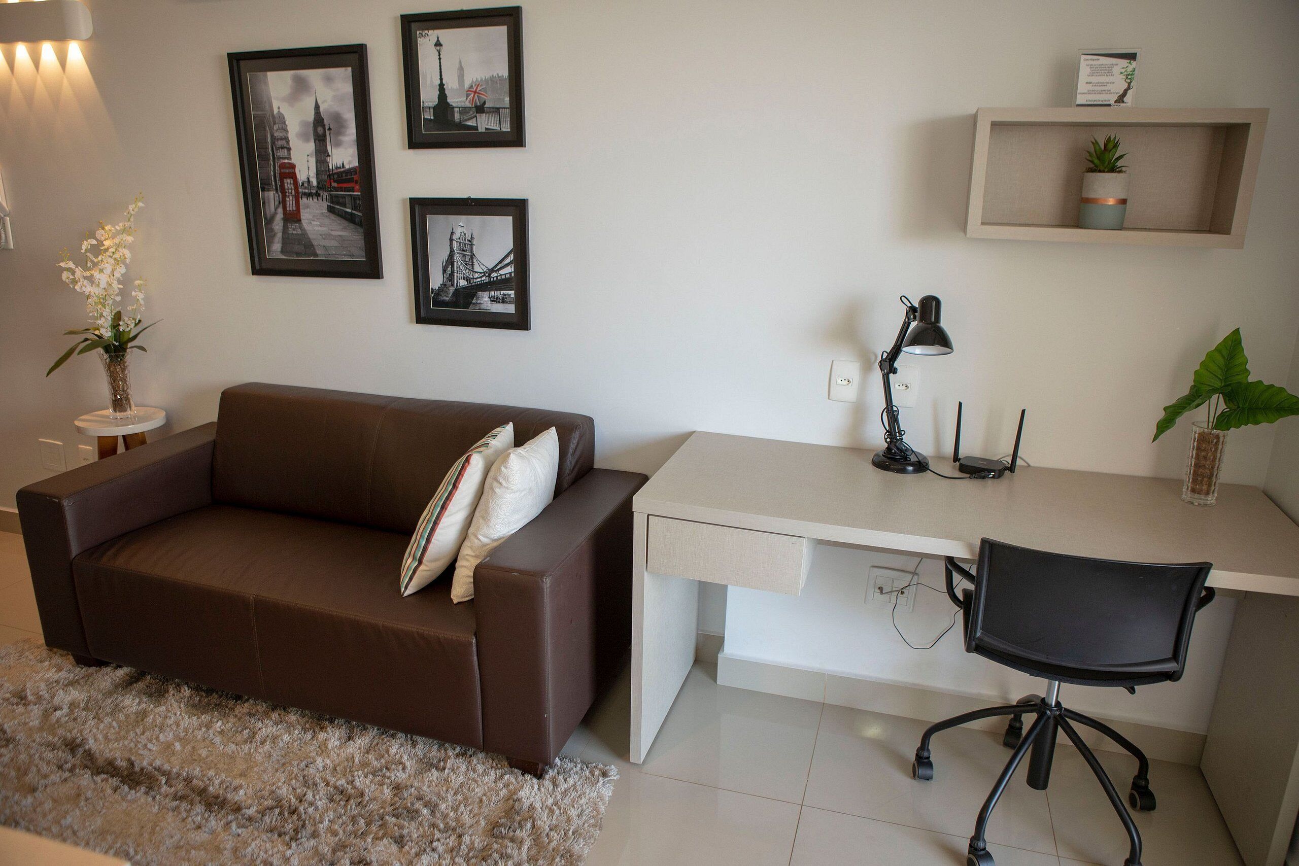 A modern room with a brown sofa, a white desk, a black office chair, a small side table with a plant, and framed pictures on the wall.