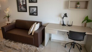 A modern room with a brown sofa, a white desk, a black office chair, a small side table with a plant, and framed pictures on the wall.