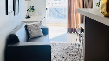 A modern living room with a sofa, a rug, a desk with a lamp, and a view of a building through the window.