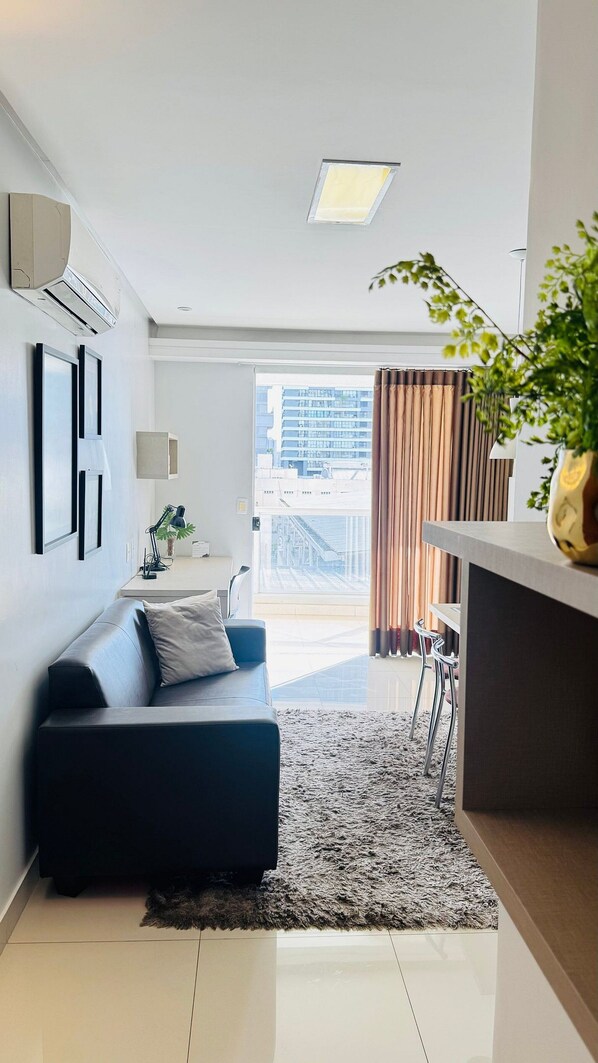 Un moderno salón con un sofá, una alfombra, un escritorio con una lámpara y vistas a un edificio a través del ventanal.