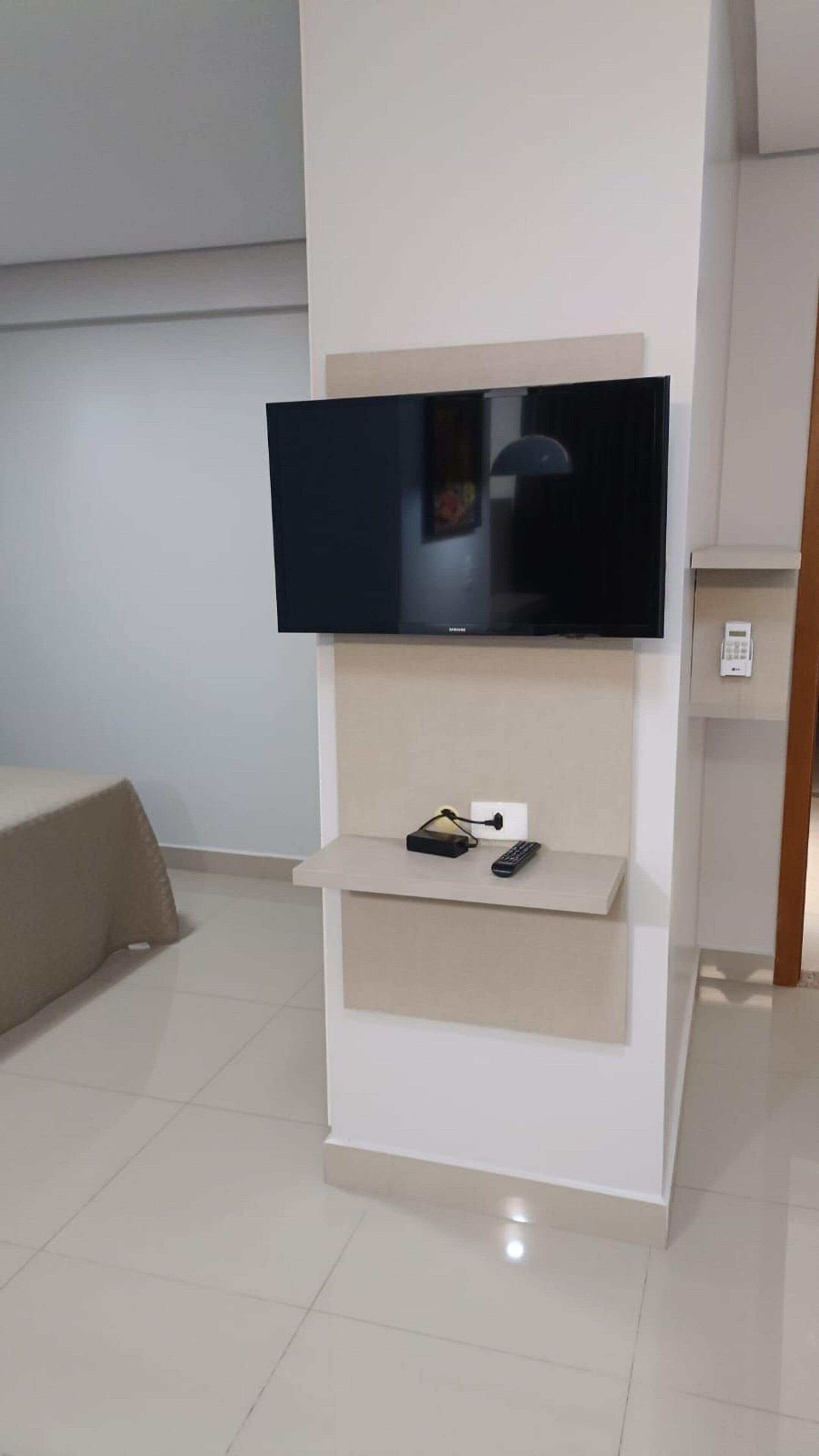 A modern living room with a flat-screen TV mounted on a white wall, a small shelf below with a remote control, and a beige sofa.