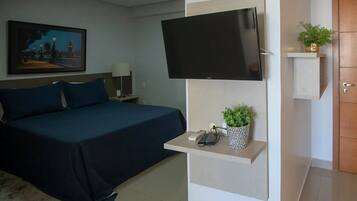 Quarto de hotel com cama, televisão montada na parede, mesa de cabeceira com telefone e controle remoto, prateleira com uma planta e porta.