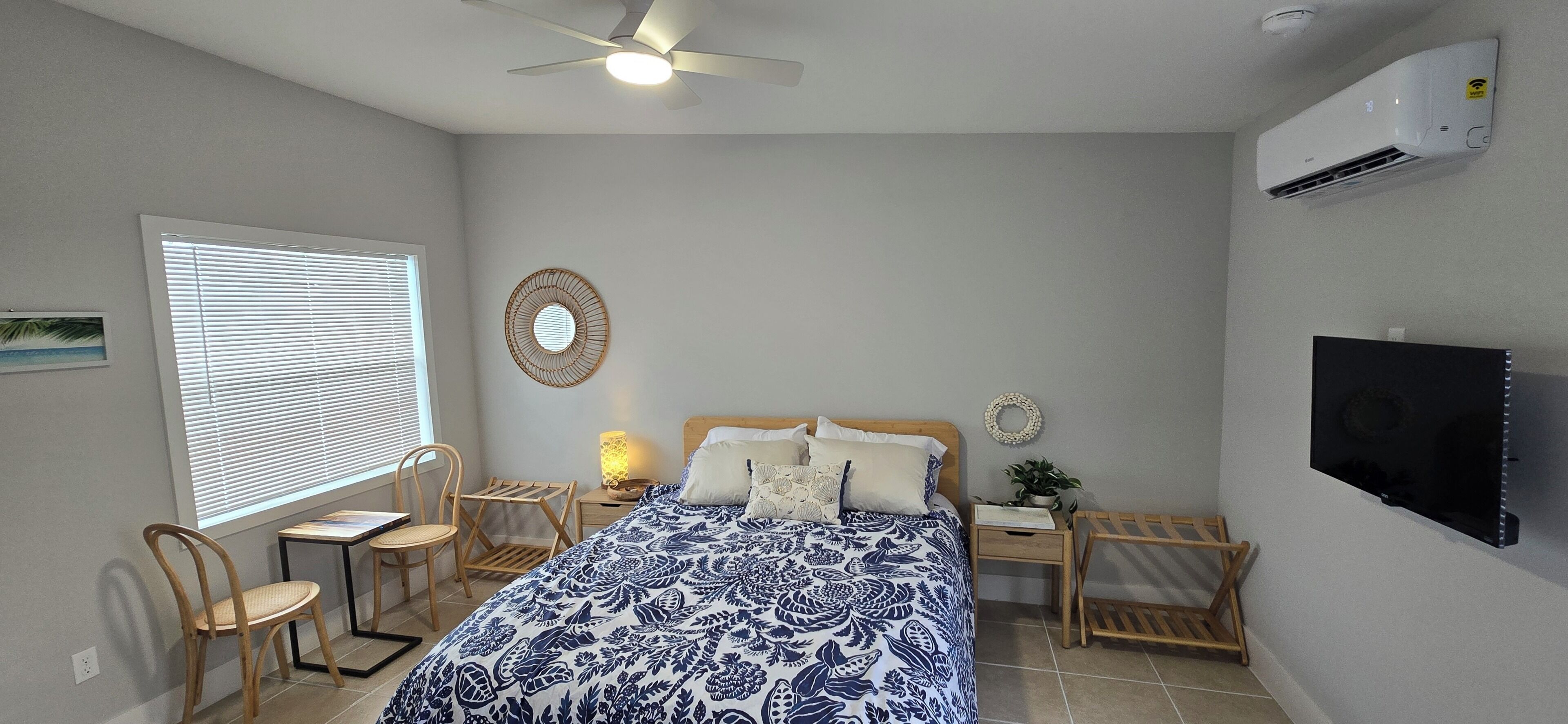 Une chambre à coucher comprenant un lit, un ventilateur de plafond, un téléviseur, une fenêtre avec des stores et un climatiseur fixé au mur.