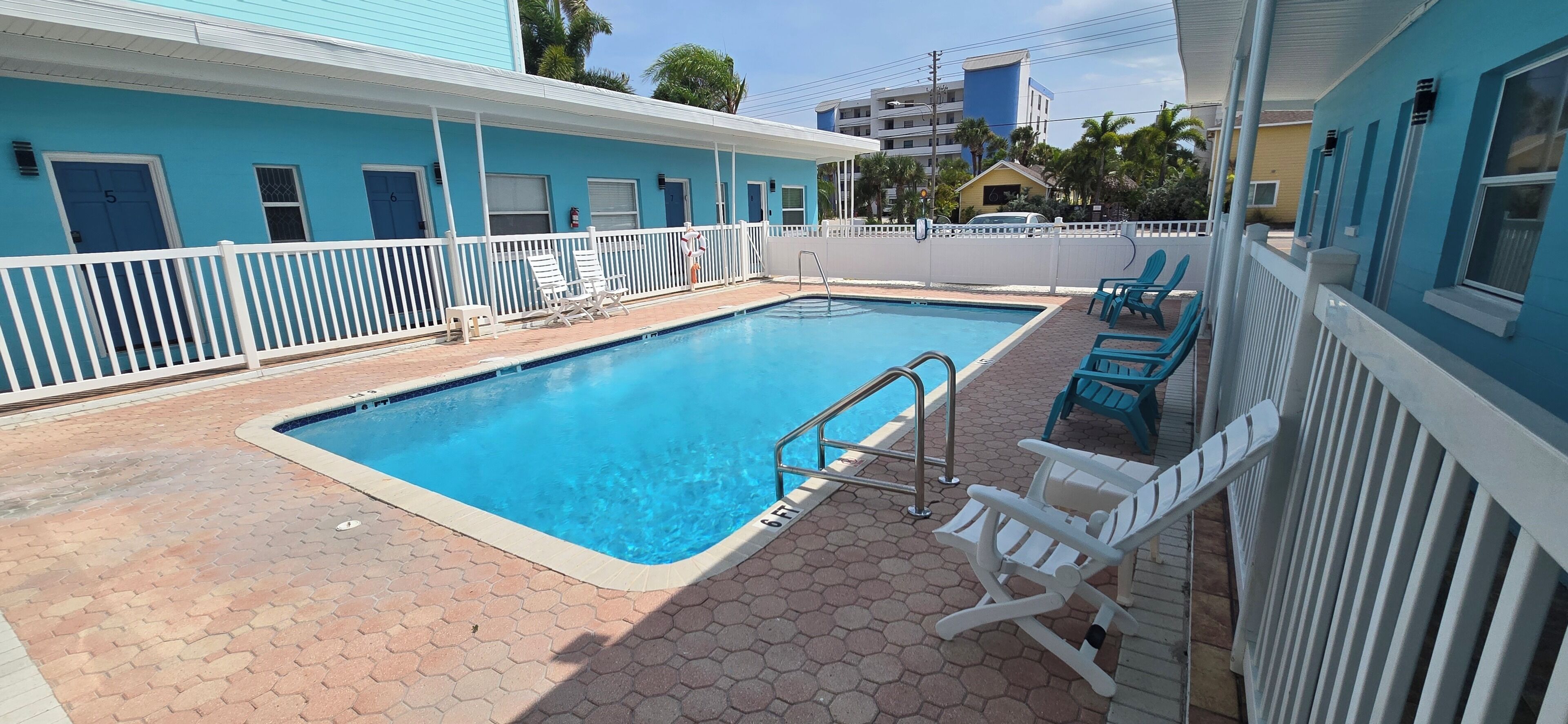 A swimming pool area with a white fence, lounge chairs, and a building with numbered doors.