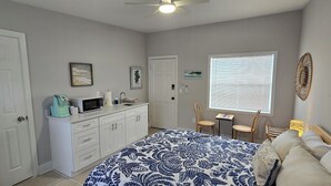 A bedroom with a bed, a desk with a microwave, a chair, a window with blinds, and a ceiling fan.
