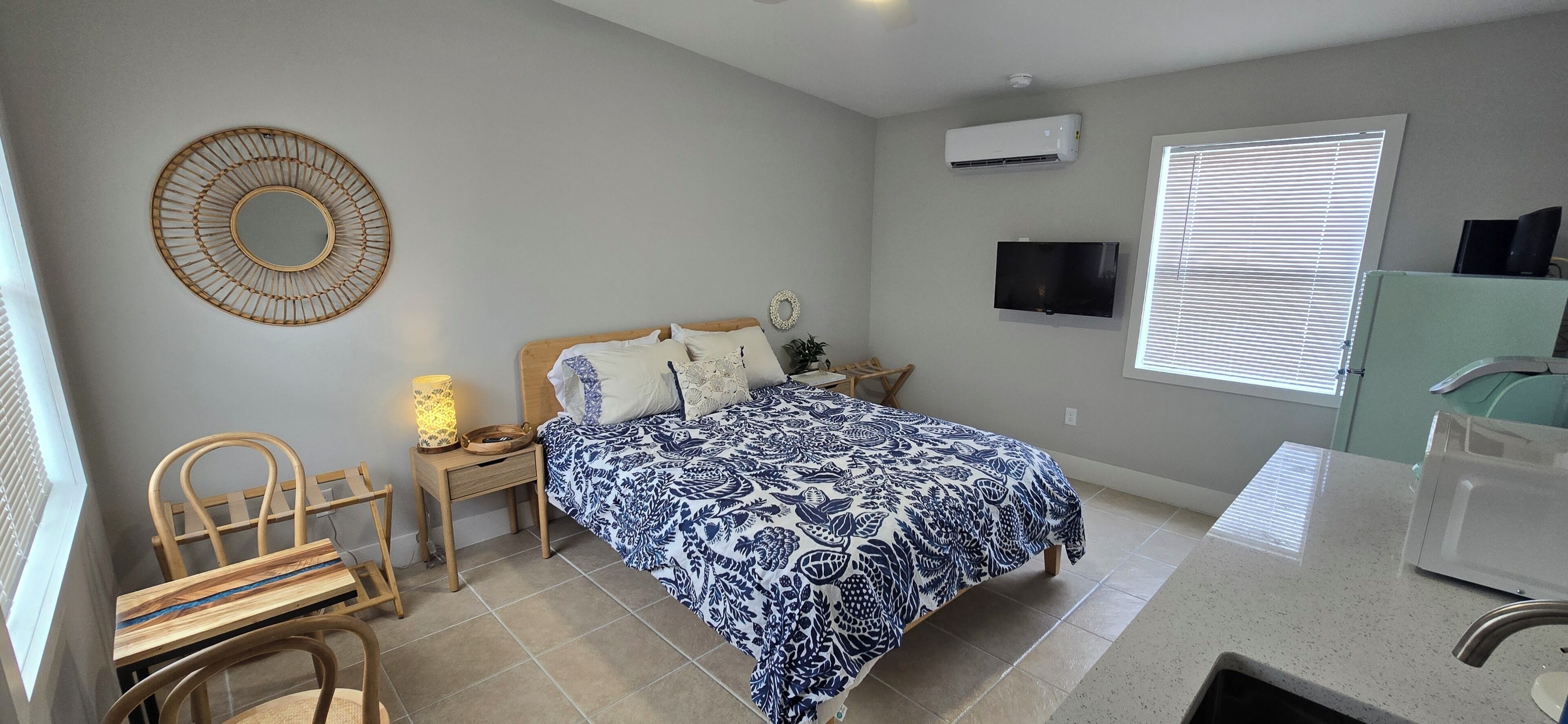A bedroom with a bed, bedside table, lamp, TV, air conditioner, and a window with blinds.