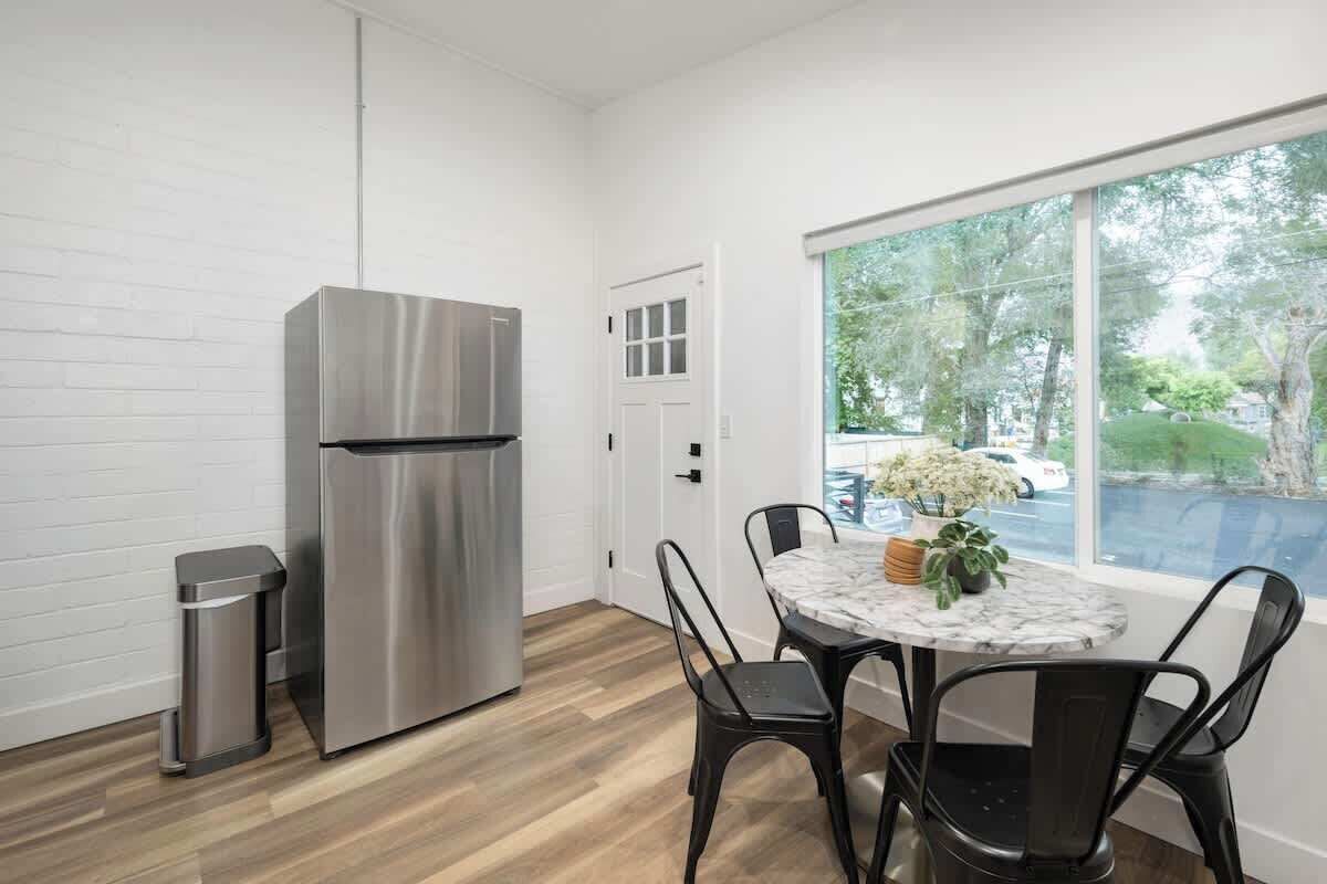 A modern kitchen with a stainless steel refrigerator, a trash bin, and a dining area with a round table and chairs.
