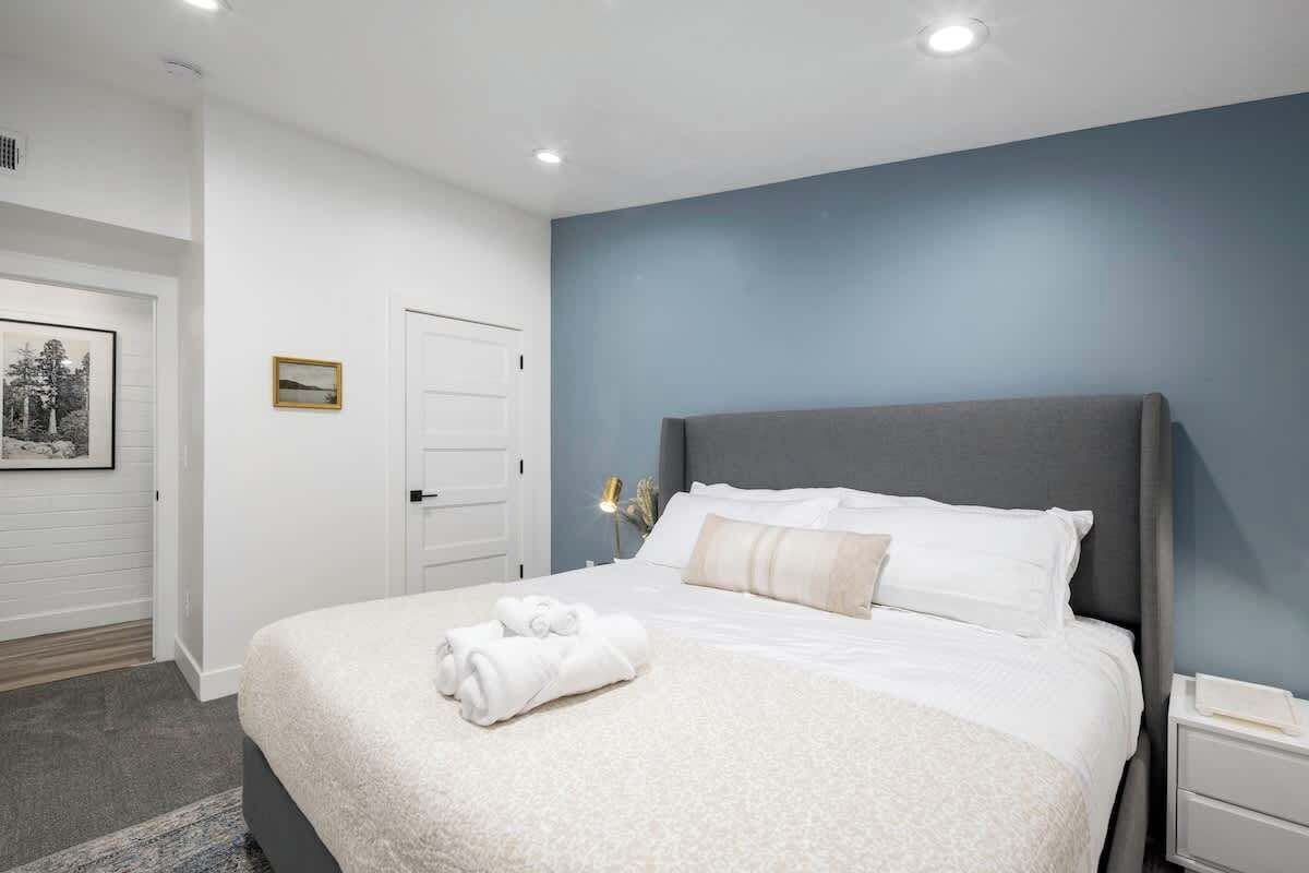 A bedroom with a large bed, white bedding, a grey headboard, a bedside table with a lamp, and a framed picture on the wall.