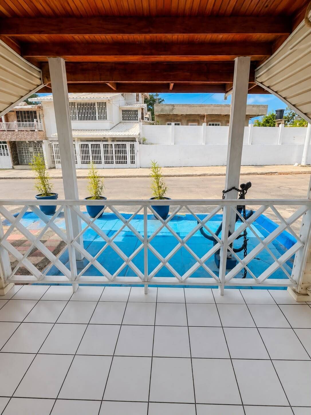 A covered balcony with a view of a swimming pool, white railings, and potted plants.