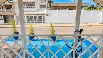 A covered balcony with a view of a swimming pool, white railings, and potted plants.