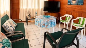 A room with a wooden ceiling, a ceiling fan, a television, a table with a tablecloth, two green chairs, and a painting on the wall. - La Casa del Ancla - A beautiful nautical cabin! (San andrés)