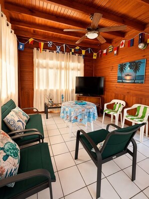 A room with a wooden ceiling, a ceiling fan, a television, a table with a tablecloth, two green chairs, and a painting on the wall. - La Casa del Ancla - A beautiful nautical cabin! (San andrés)