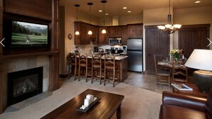 A spacious living room with a fireplace, wooden furniture, a television, and a kitchen area with modern appliances and pendant lighting. - Luxury 2BR-Sleeps 8! Near Gondola and Hiking-Hot Tubs, Spa, A/C-June 2027 (Breckenridge)