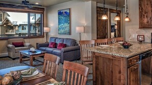 A spacious living area with a large window, a dining table with chairs, a kitchen with wooden cabinets, and a ceiling fan. - Luxury 2BR-Sleeps 8! Near Gondola and Hiking-Hot Tubs, Spa, A/C-June 2027 (Breckenridge)