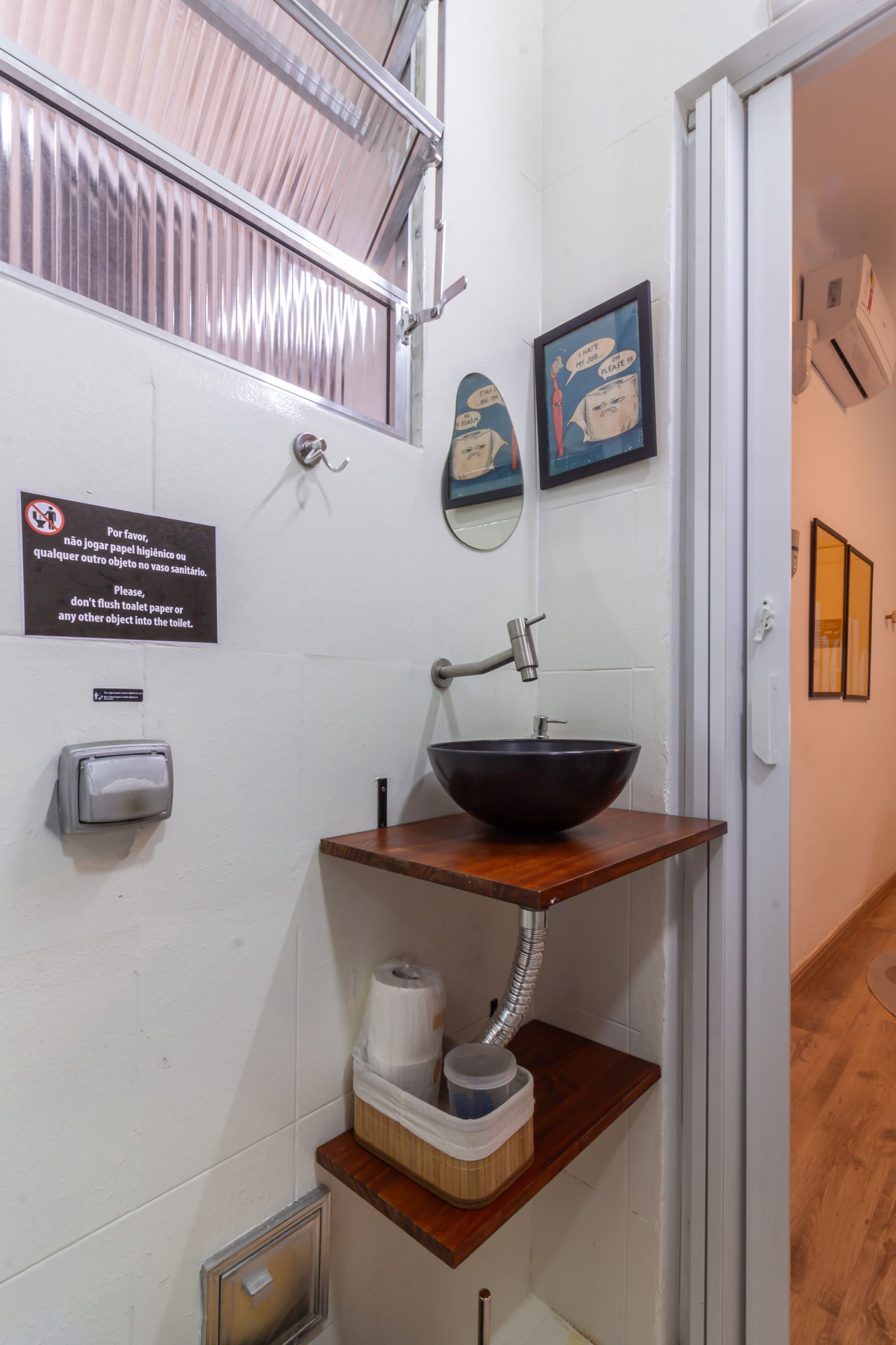 Un baño con lavamanos, papel higiénico, secamanos automático y un cubo de basura.