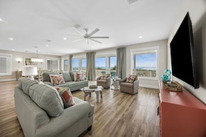 A spacious living room with a large TV, a ceiling fan, and a view of the ocean.