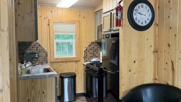 A compact kitchen with wooden walls, a stainless steel sink, a black refrigerator, and a clock on the wall.