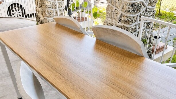 Una mesa de madera con estructura metálica, sobre un suelo de concreto con barandilla blanca y plantas en macetas al fondo.