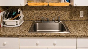 Cocina con encimera de granito, pileta de acero inoxidable, gabinetes blancos y un escurreplatos con utensilios.
