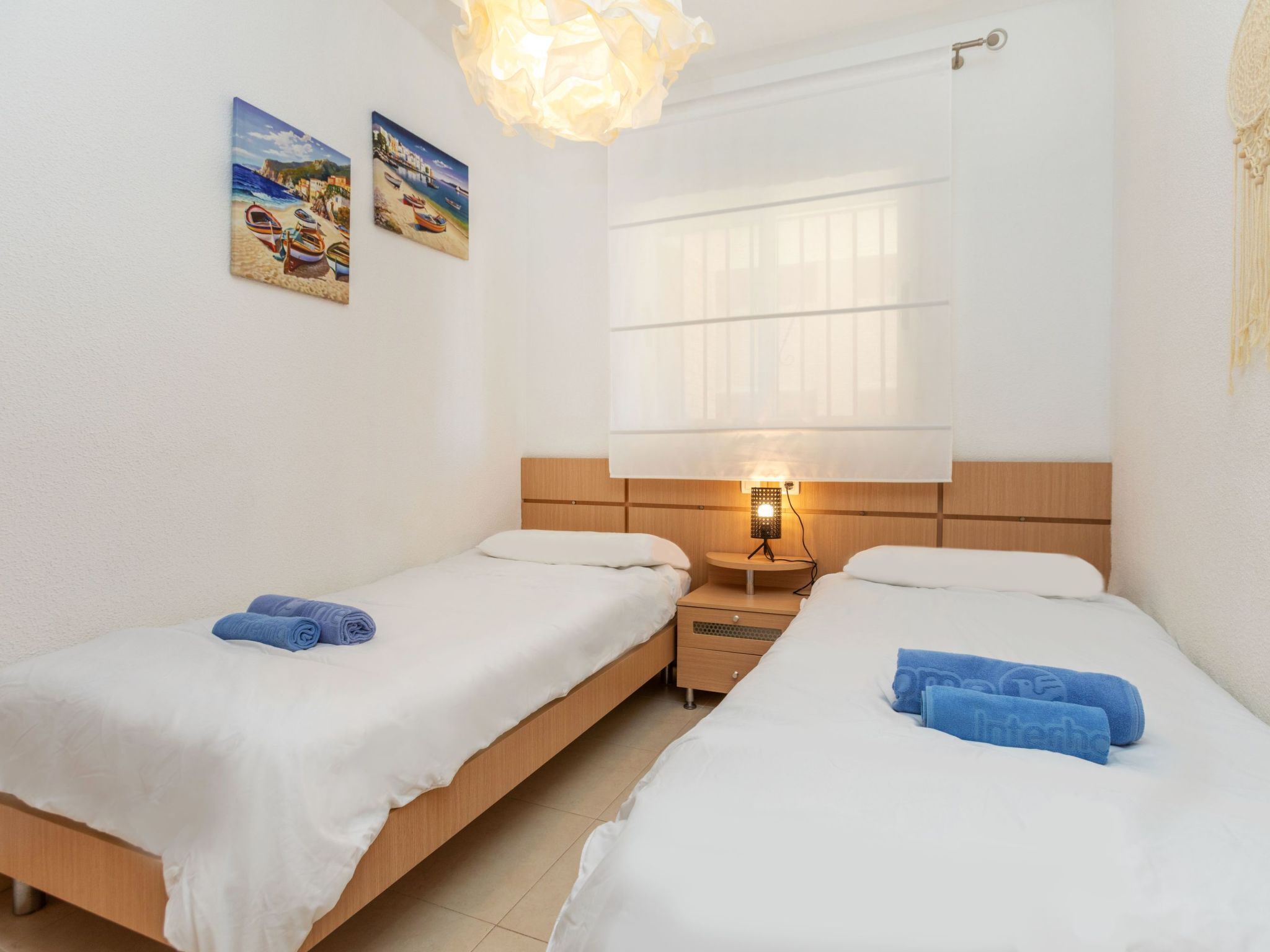 A hotel room with two single beds, a nightstand with a lamp, a window with curtains, and two wall art pieces depicting coastal scenes.