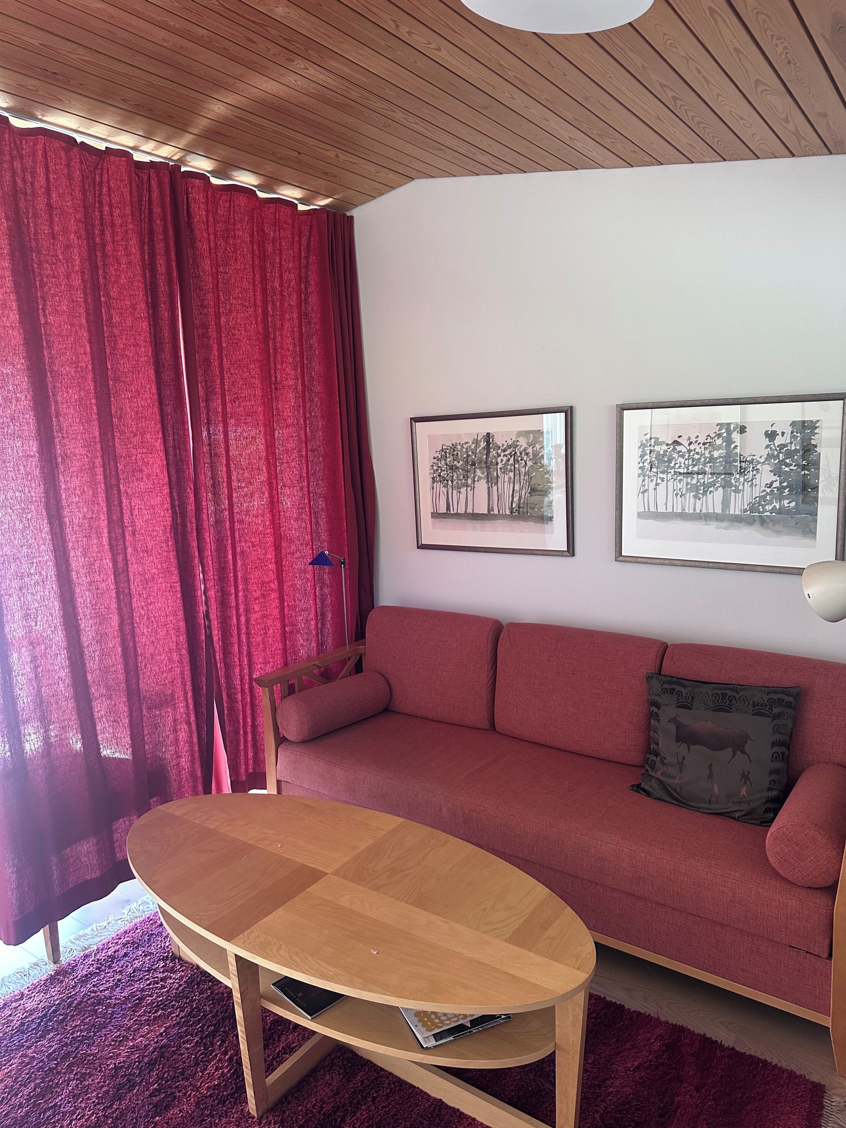Een woonkamer met een houten plafond, een rode bank, een houten salontafel en twee ingelijste foto's aan de muur.
