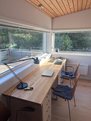 Un espace de bureau moderne avec un bureau en bois, deux chaises, un ordinateur portable, une lampe de bureau et de grandes fenêtres donnant sur un paysage verdoyant.
