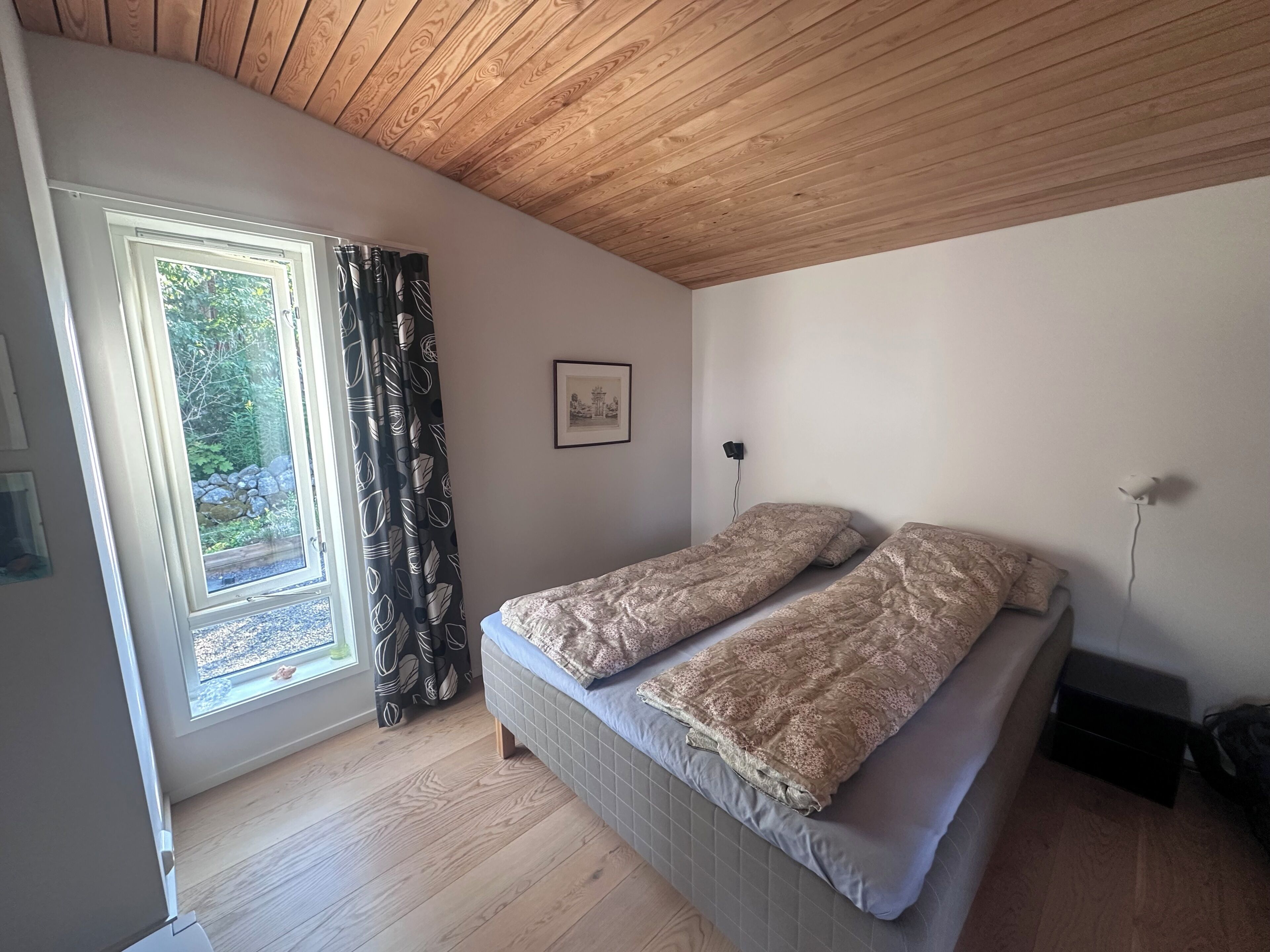 Ein Schlafzimmer mit Holzbalkendecke, einem Bett mit gemusterten Betttextilien, einem Fenster mit Vorhängen und einem kleinen Bild an der Wand.