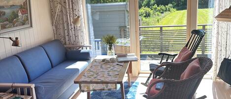A cozy living room with a blue sofa, wicker chairs, a wooden table, and a view of a garden through large windows.
