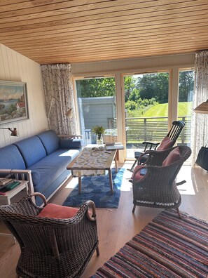 Un salon chaleureux avec un canapé bleu, des fauteuils en osier, une table en bois et une vue sur un jardin, visible à travers de grandes fenêtres.