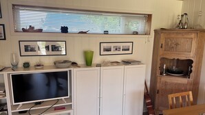 A compact living space with a television, storage cabinets, a dining table, and a window with blinds. - A large cabin on the outskirts of Tromøya, with Raet National Park as its closest neighbor. (Agder)